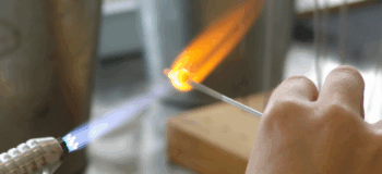 Eine Hand hält ein Stäbchen, auf dem eine Glasperle steckt, in eine Flamme eines Gasbrenners.