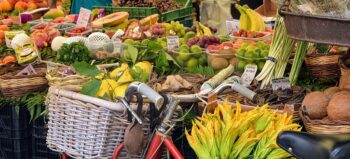 buntes Obst und Gemüse an einem Marktstand. Davor lehnt ein rotes Fahrrad. In einem Korb: Zucchiniblüten.