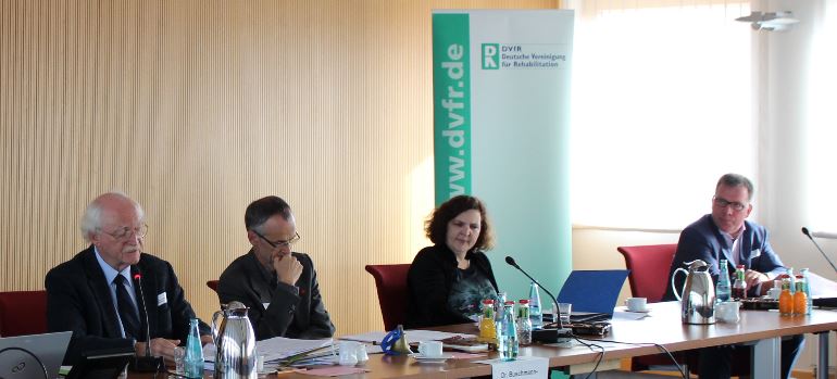 Vier Menschen an einem Konferenztisch, dahinter ein Banner der Deutschen Vereinigung für Rehabiliation e.V. Heidelberg.