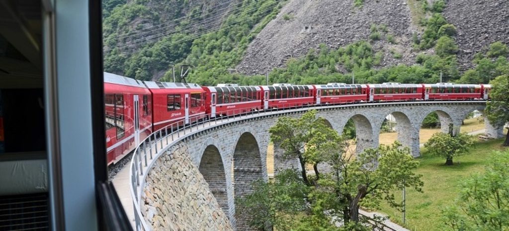 Roter Zug fährt über steinerne Bogenbrücke durch Tal.
