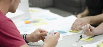 Mehrere Menschen (einer im Anschnitt zu sehen, von den anderen sind nur die Hände zu sehen) sitzen an einem Tisch bei Planungsarbeiten mit bunten Post-Its.