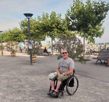 Mann in Rollstuhl vor Hafenpromenade mit Oleanderbüschen und Bäumen.