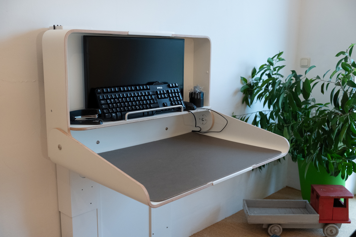 Aufgeklappter und umfunktionierter Wickeltisch, der nun als Computerarbeitsplatz und Schreibtisch dient. An der Wandseite sind ein Bildschirm, eine Tastatur und Büromaterialien gelagert.