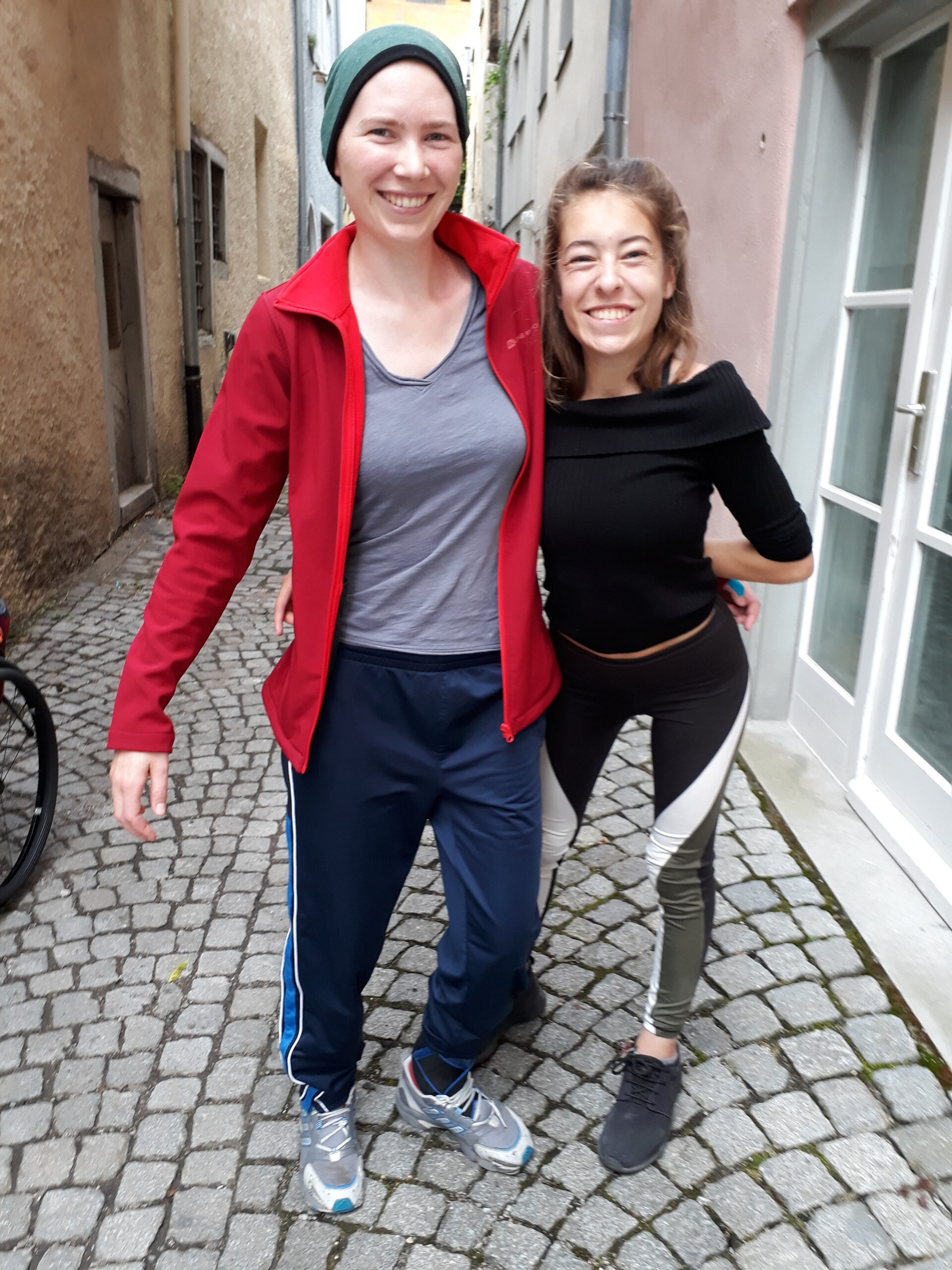 Zwei junge Frauen stehen in einer schmalen Kopfsteinpflaster-Gasse, umarmen sich und lachen in die Kamera.