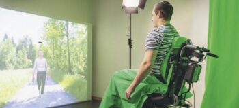 Mann im Elektrorollstuhl sitzt vor Leinwand, auf die ein Film mit einem gehenden Mann projiziert wird.
