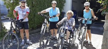 Brigitte Loos im Handbike. Drei ihrer Freundinnen stehen mit Rädern und im Fahrraddress neben ihr im strahlenden Sonnenschein vor einem geöffneten Garagentor.