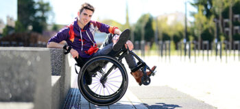 Junger Rollstuhlfahrer (Moritz Brückner) in violetter Jacke mit orangenen Verzierungen, Jeaens und braunen Stiefeletten lehnt in einem sonnigen Park mit aufgestütztem Ellenbogen an einer Betonbrüstung.