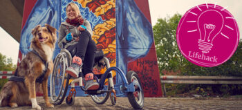 Eine junge blonde Frau im Rollstuhl vor einer bunten Wand. Links von ihr ein Hund. Rechts von ihr der rosa Button mit einer Glühbirne und der Aufschrift Lifehack.