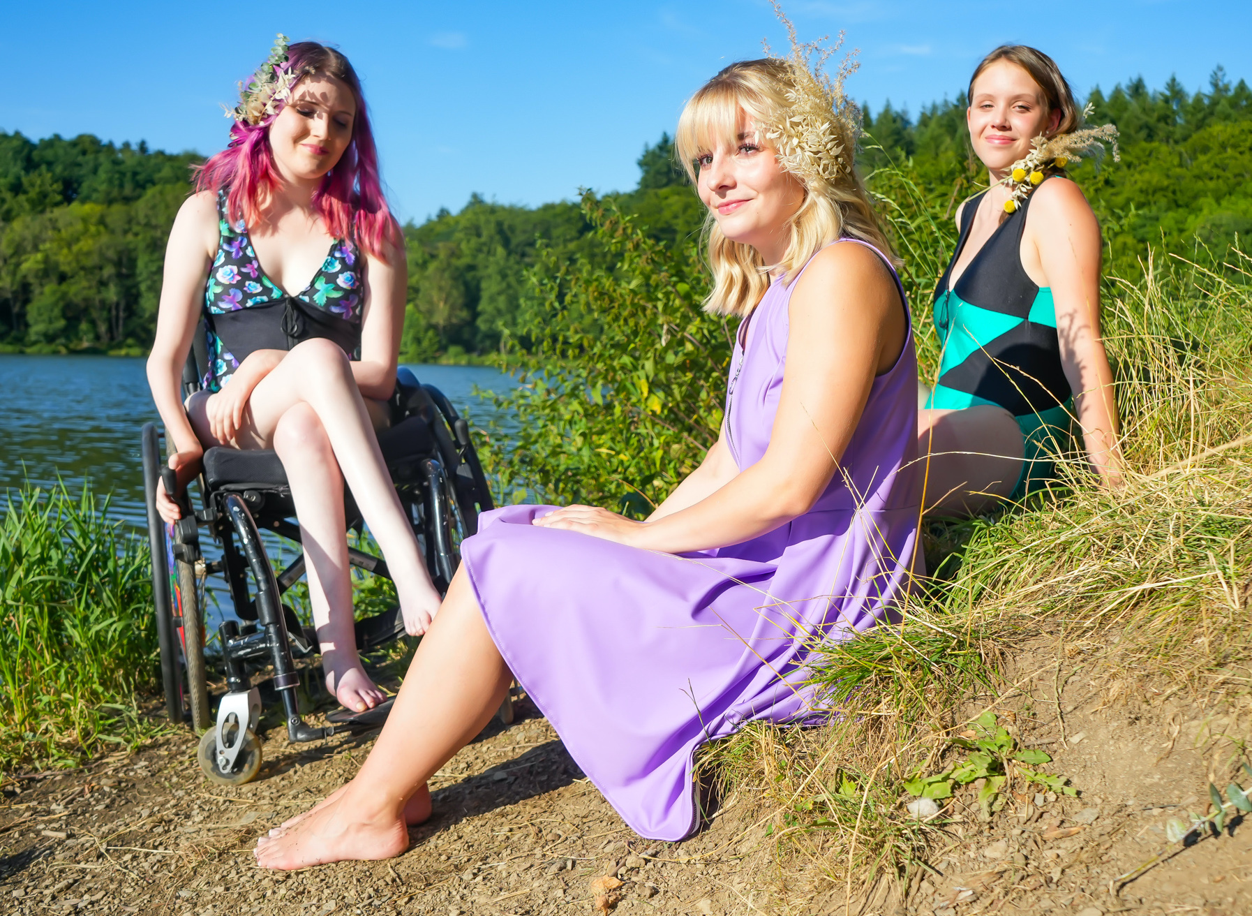 3 junge Frauen in bunter Freizeit- und Badekleidung an einem See. Zwei von ihnen (eine davon im Rollstuhl) tragen Bademode. Ihre Haare haben sie mit Blumen und Gräsern geschmückt.