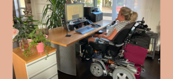 Steffen in ihrem E-Rolli mit Kinnsteuerung vor ihrem Schreibtisch, auf dem Geräte (Smartphone, Computer) stehen.
