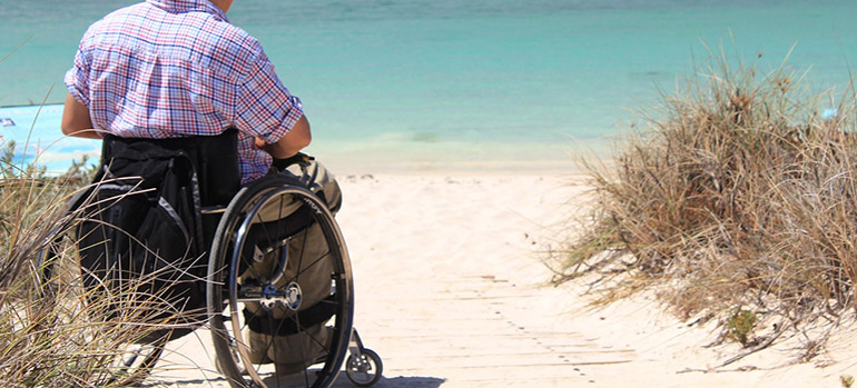 Mann in kariertem Hemd und im Rollstuhl am Strand. Im Hintergrund ein Gewässer.