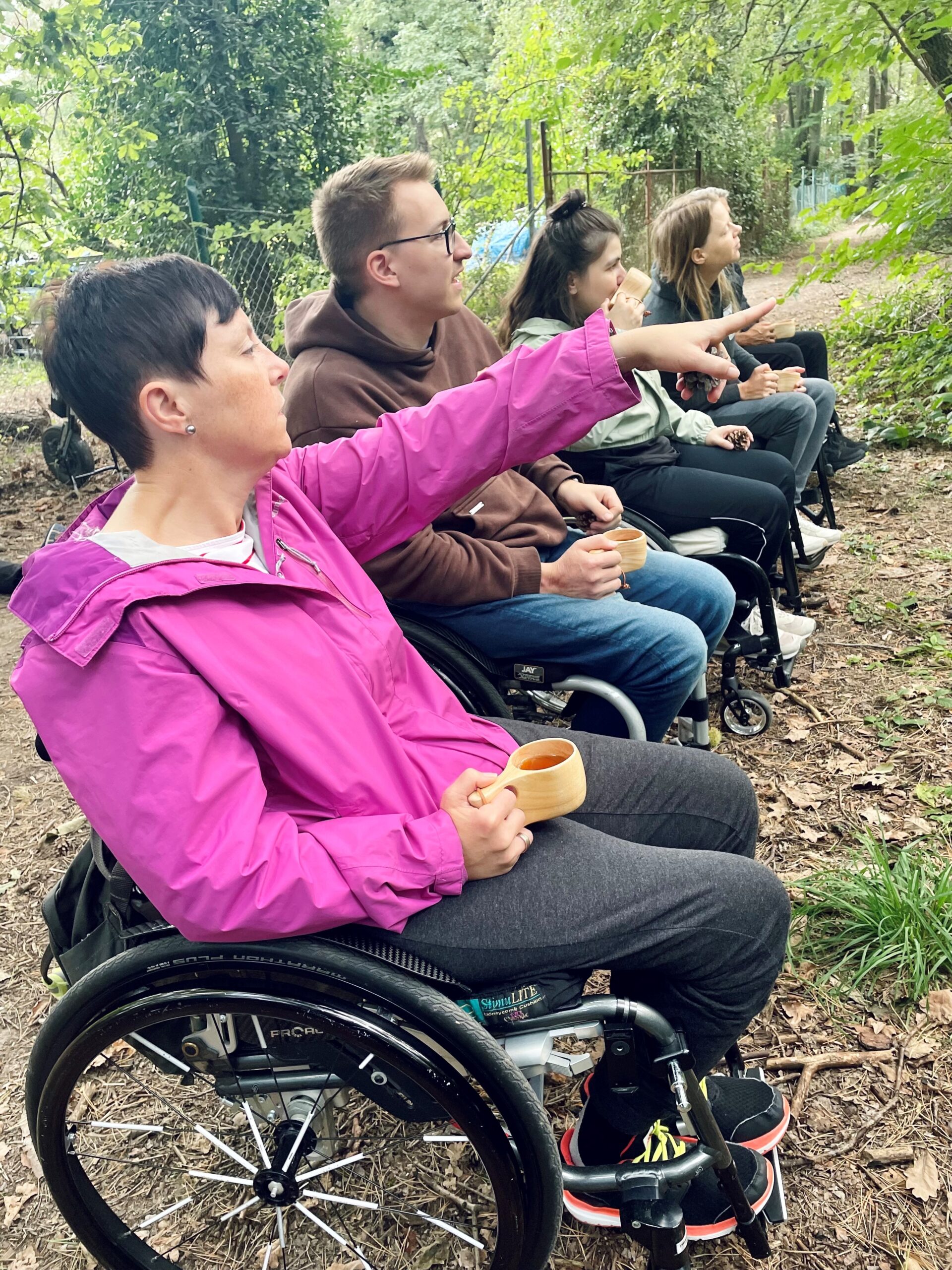 Fünf Menschen im Rollstuhl blicken in die Natur, eine deutet auf etwas. In den Händen halten sie Tassen aus Holz.