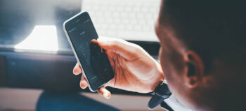 Mann an Schreibtisch hält Smartphone in einer Hand.