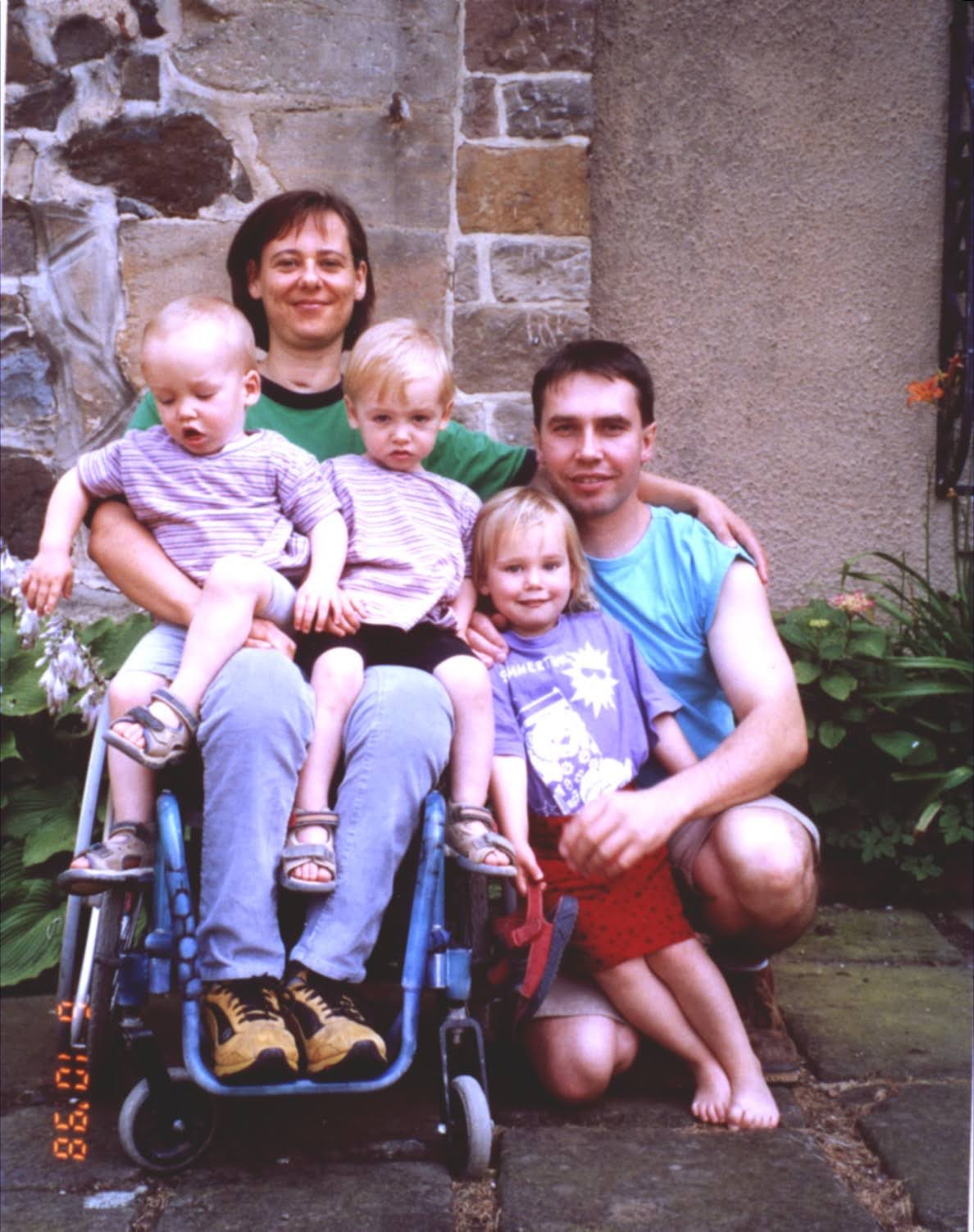 Familienfoto. Die Mutter im Rollstuhl hat zwei Kinder auf dem Schoß, der Vater hält das größere Kind umarmt.