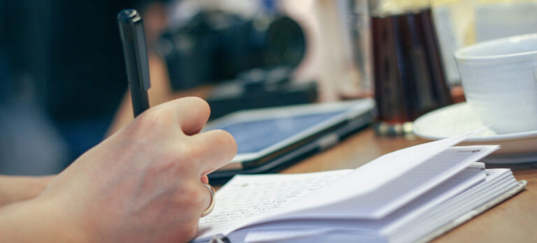 Hand mit Ehering hält Kugelschreiber, schreibt Notizen in ein Buch. Auf dem Schreibtisch liegen zudem ein Tablet, eine Kamera, neben dem Buch steht eine Tasse und eine Karaffe.