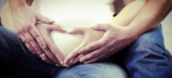 Zwei paar Hände umschließen den Babybauch einer Frau.