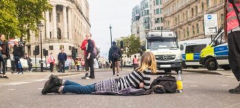 Blonde Frau in Jeans und Ringelshirt liegt auf der Straße. Menschen gehen und stehen auf der Straße. Im Hintergrund: 3 Polizeiwagen.