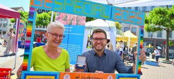 Eine lächelnde Frau mit blonden Haaren, Brille und grünem Shirt und ein lächelnder Mann mit dunklen Haaren, Bart, Brille und blauem Shirt halten einen Rahmen aus bunten Legosteinen vor sich. Die Aufschrift: Be Special 2025