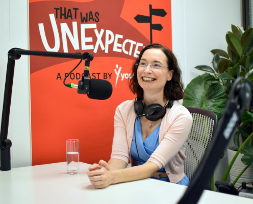 Frau in blauem Kleid und rosa Strickjacke sitzt an weißem Schreibtisch, vor sich ein großes Podcast-Mikrofon und lächelt, einen Kopfhörer um den Nacken gelegt. Hinter ihr ein orangenes Plakat mit der Aufschrift „Theat was unexpectable“.