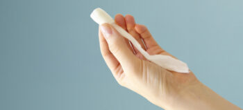 Frauenhand hält vor hellblauem Hintergrund einen Analtampon aus weißem Schaumstoffgewebe.