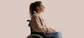 Junge Frau mit Pferdeschwanz, rose-farbenem Strickpulli und Jeans sitzt in einem Rollstuhl und lächelt zart.