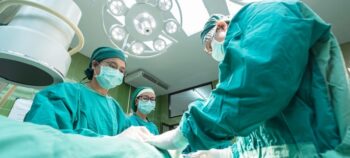 Drei medizinische Fachpersonen in grüner OP-Bekleidung und Mundschutz stehen in einem OP-Saal.