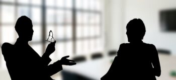 Ein Konferenzraum mit hohen Fenstern. Im Vordergrund die schemenhaften Figuren von zwei sitzenden Menschen.