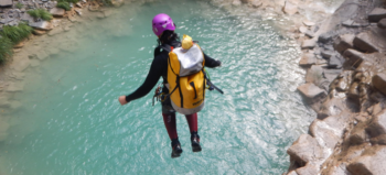 Symbolbild: Mensch in Sportmontur, mit Helm und großem gelben Rucksack springt aus relativ großer Höhe in einen Bergsee.