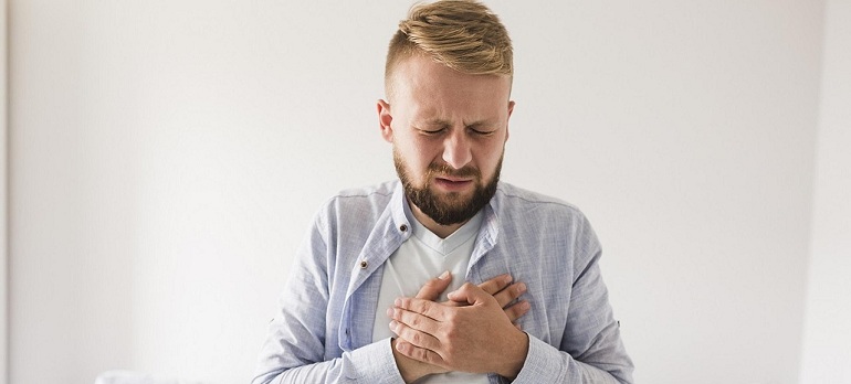 Blonder Mann mit Bart in hellen Kleidern und mit schmerzverzerrtem Gesicht hält sich beide Hände an die Brust.