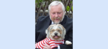 Mann in schwarzem Anzug hält kleinen Hund auf dem Schoss. Der Hund trägt ein weiß-rotes Ringel-T-Shirt.