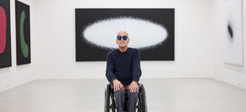 Wolfgang Temmel im Rollstuhl in einem Ausstellungsraum. Er ist schwarz gekleidet, trägt eine Sonnenbrille. Hinter ihm sein Gemälde "Ein Lichtjahr".