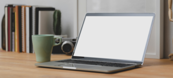 Aufgeschlagener Laptop mit leerem Bildschirm auf Holztisch, daneben eine grüne Tasse, ein Kaktus, eine Fotoapparat und aufgereihte Notiz-Bücher.