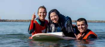 Eine Surflehrerin, ein Surflehrer im Meer, in orangefarbenen Schwimmwesten, zwischen ihnen: Eine junge Frau, die auf einem Surfbrett liegt.
