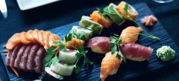 Sushi von Lachs und Tunfisch arrangiert auf einer schwarzen Schieferplatte dekoriert mit Korianderblättern. Daneben ein Klecks Wasabi.
