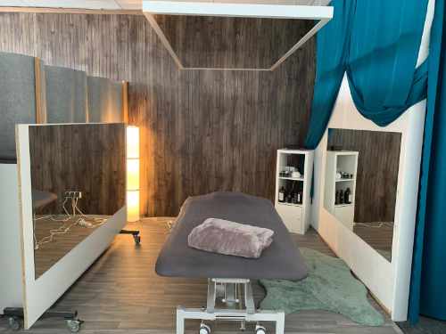 Ein Behandlungsraum mit Ganzkörperspiegeln an zwei Seiten und der Decke. Die Behandlungsliege hat einen grauen Bezug; darauf liegt eine gefaltete graue Decke. Der Raum ist mit gelbem Licht sanft beleuchtet.