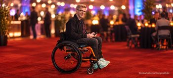 Mann mit Brille sitzt in einem Rollstuhl mit orangefarbenem Rahmen auf einem roten Teppich bei einer festlichen Abendveranstaltung. Im unscharfen Hintergrund sind Gäste, Stehtische und warme Lichter zu sehen.